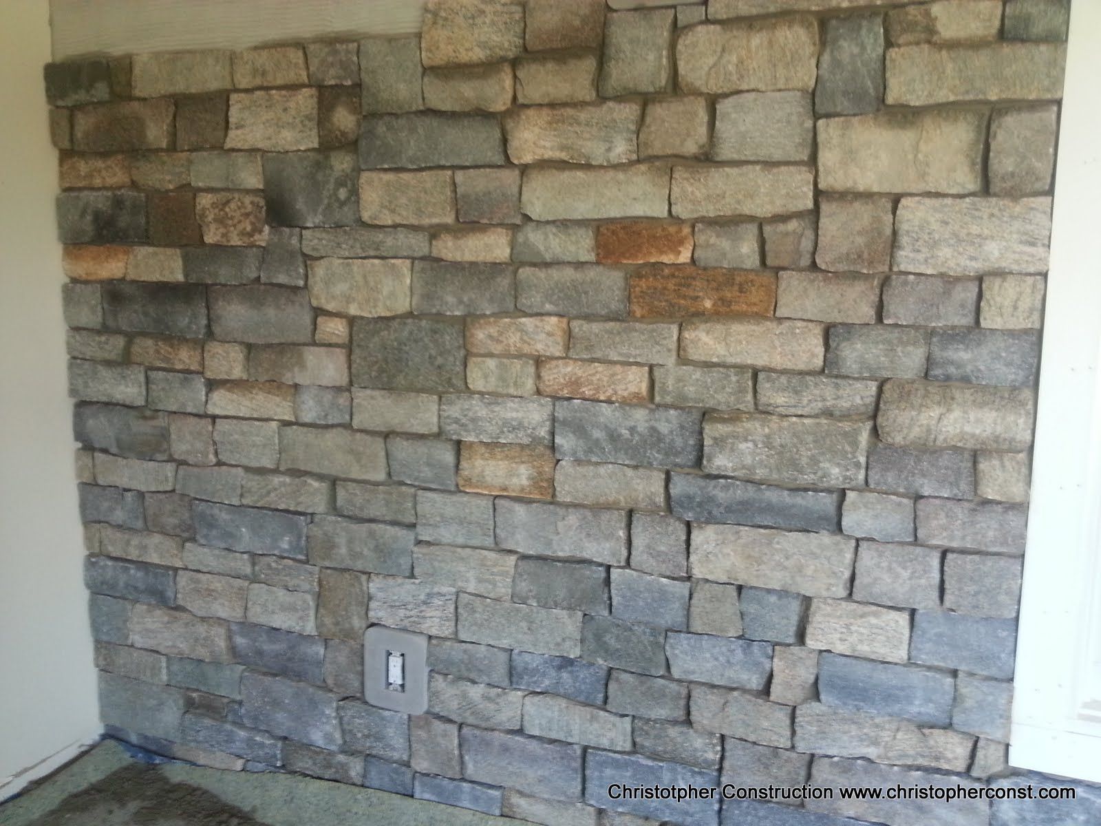 Stone accent wall with gray and tan stacked stone tiles in a room corner, with an electrical outlet near the bottom