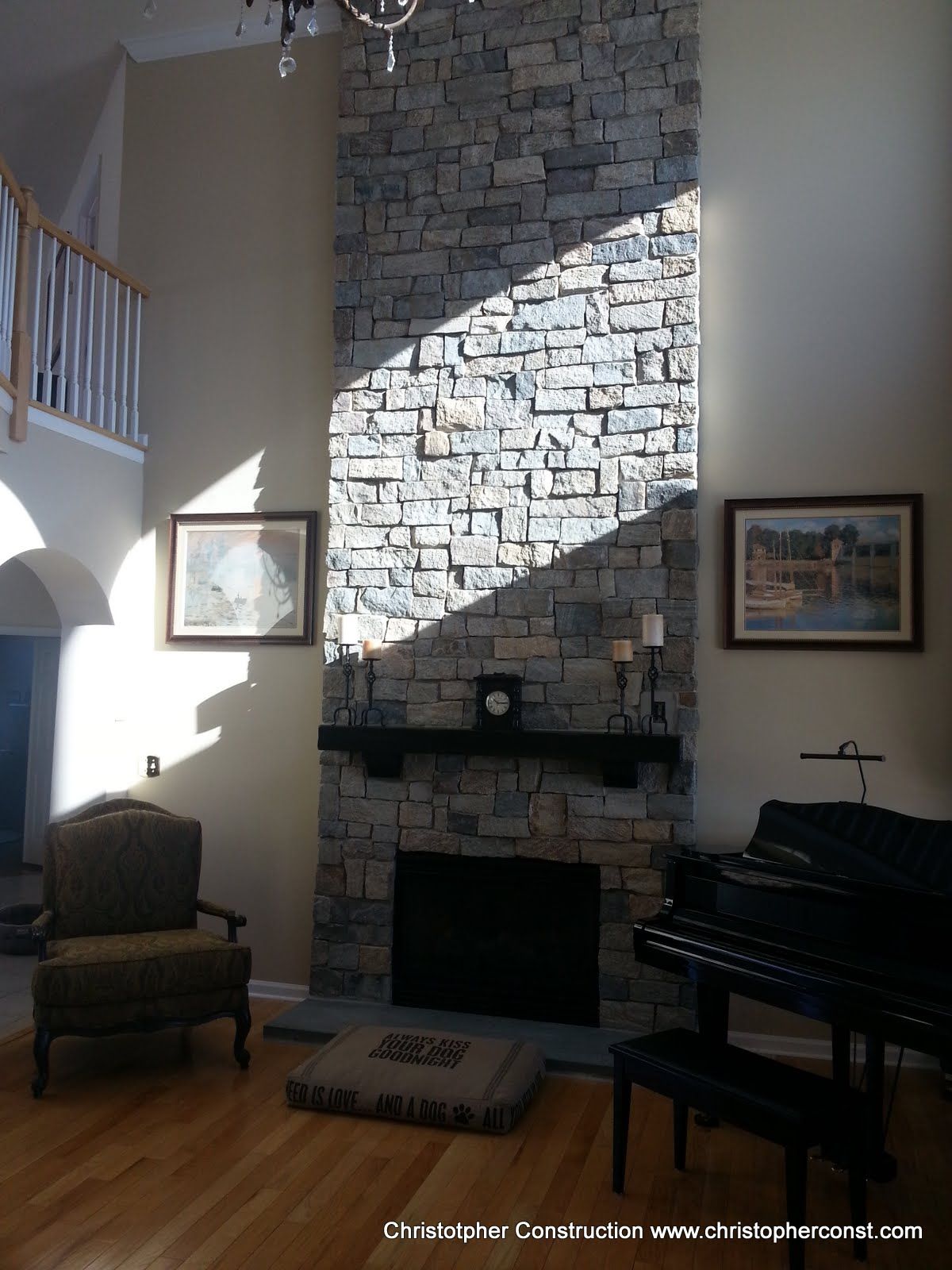Living room with stone fireplace, piano, armchair, and staircase in warm sunlight.