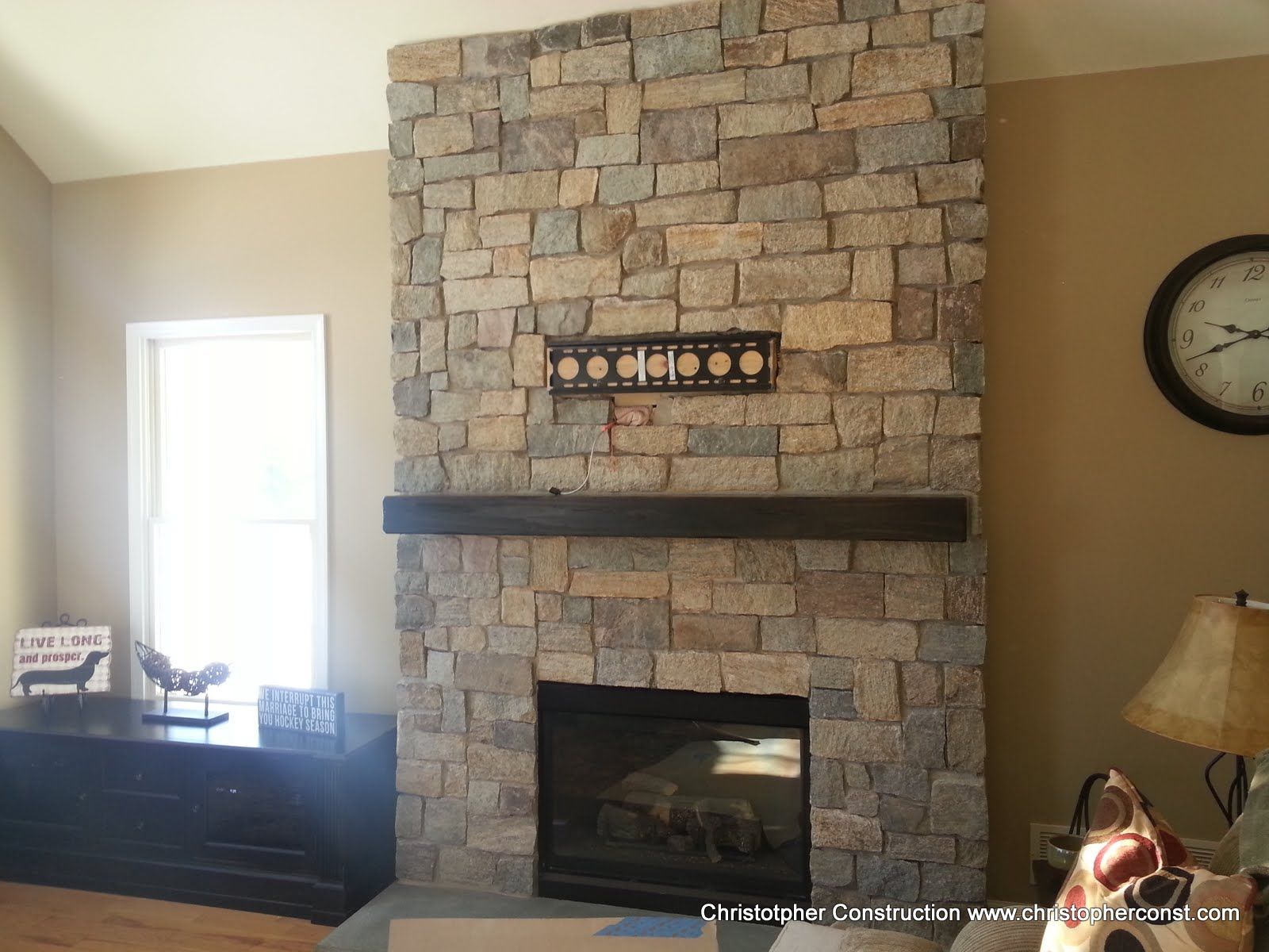 Living room with stone fireplace, dark mantel, and wall clock beside a window and sideboard
