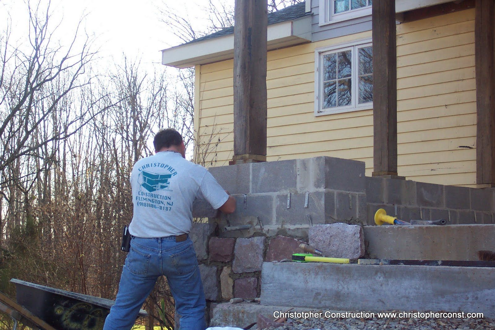 Worker stacking cinder blocks to build a retaining wall beside a yellow house