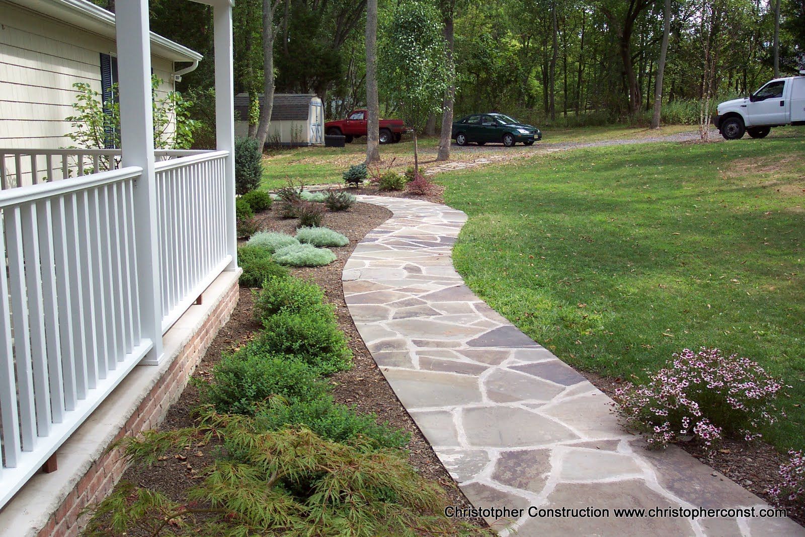 Stone garden path curving past a white porch rail through landscaped yard with trees and parked vehicles