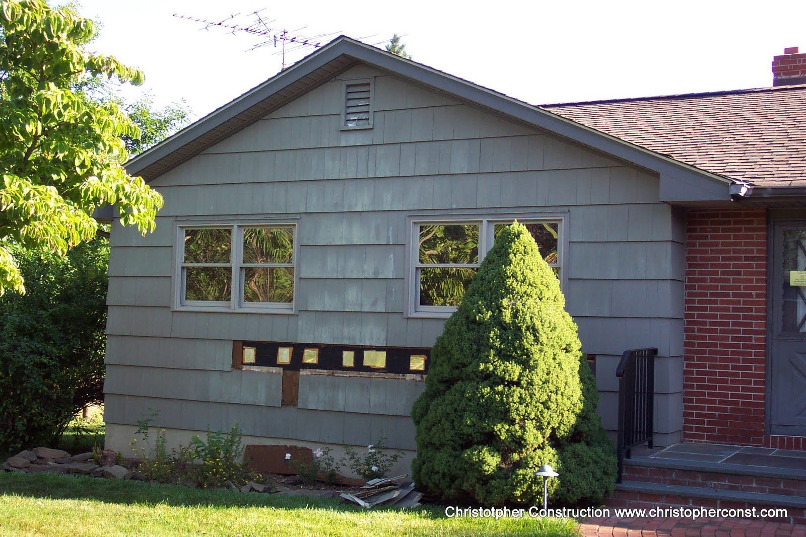 Suburban house exterior with gray siding, brick facade, and a small evergreen shrub in front.