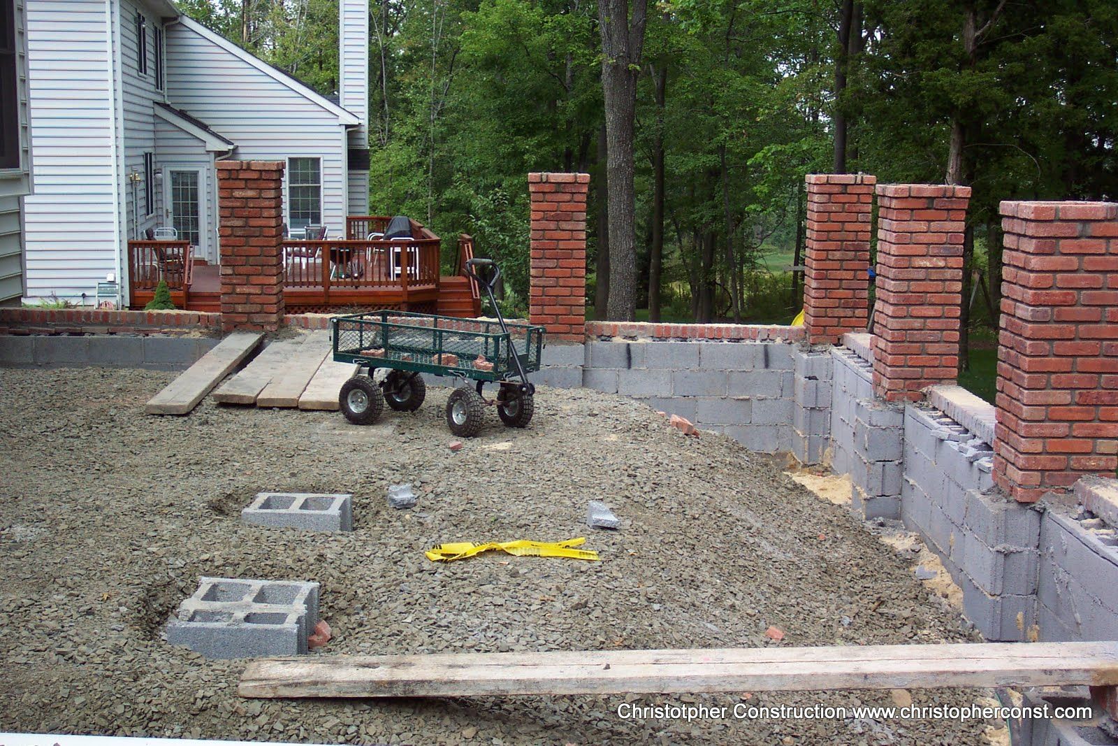 Backyard construction site with gravel foundation, brick pillars, and a wheelbarrow near a house