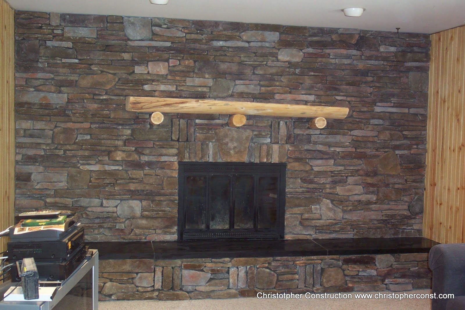 Stone fireplace with wooden mantel in a rustic room, flanked by wood paneling and a low dark hearth.