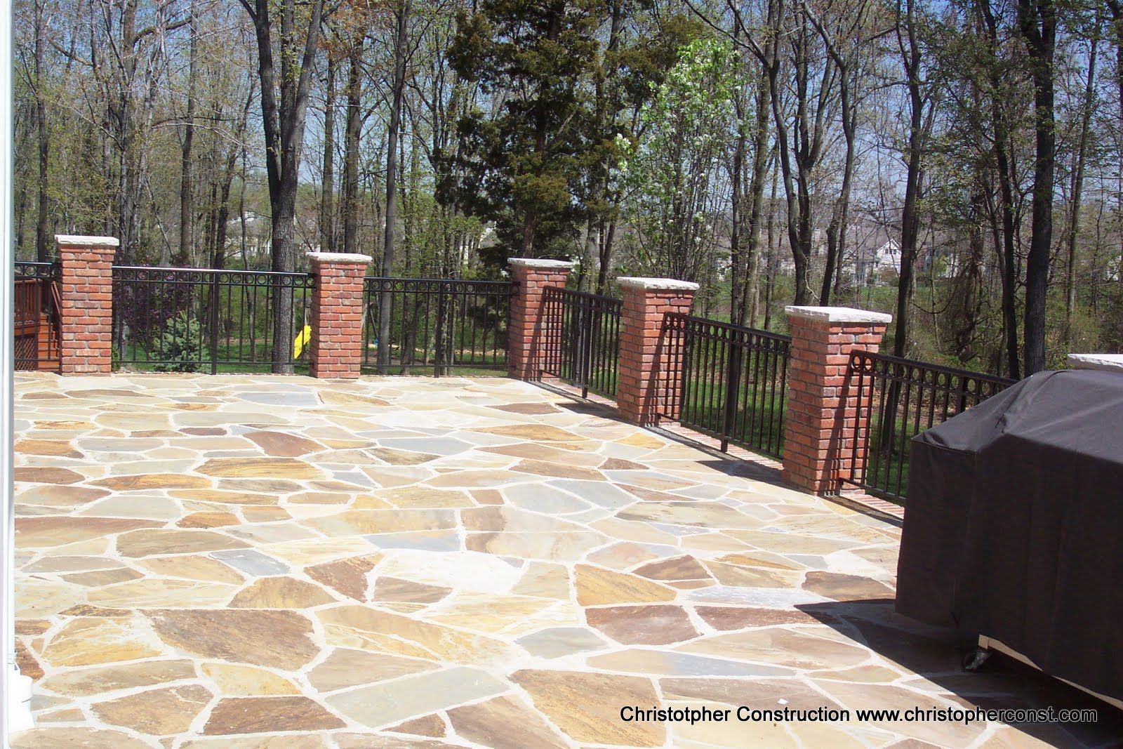 Stone patio with brick posts and black railings beside a wooded backyard, with a covered grill on the right