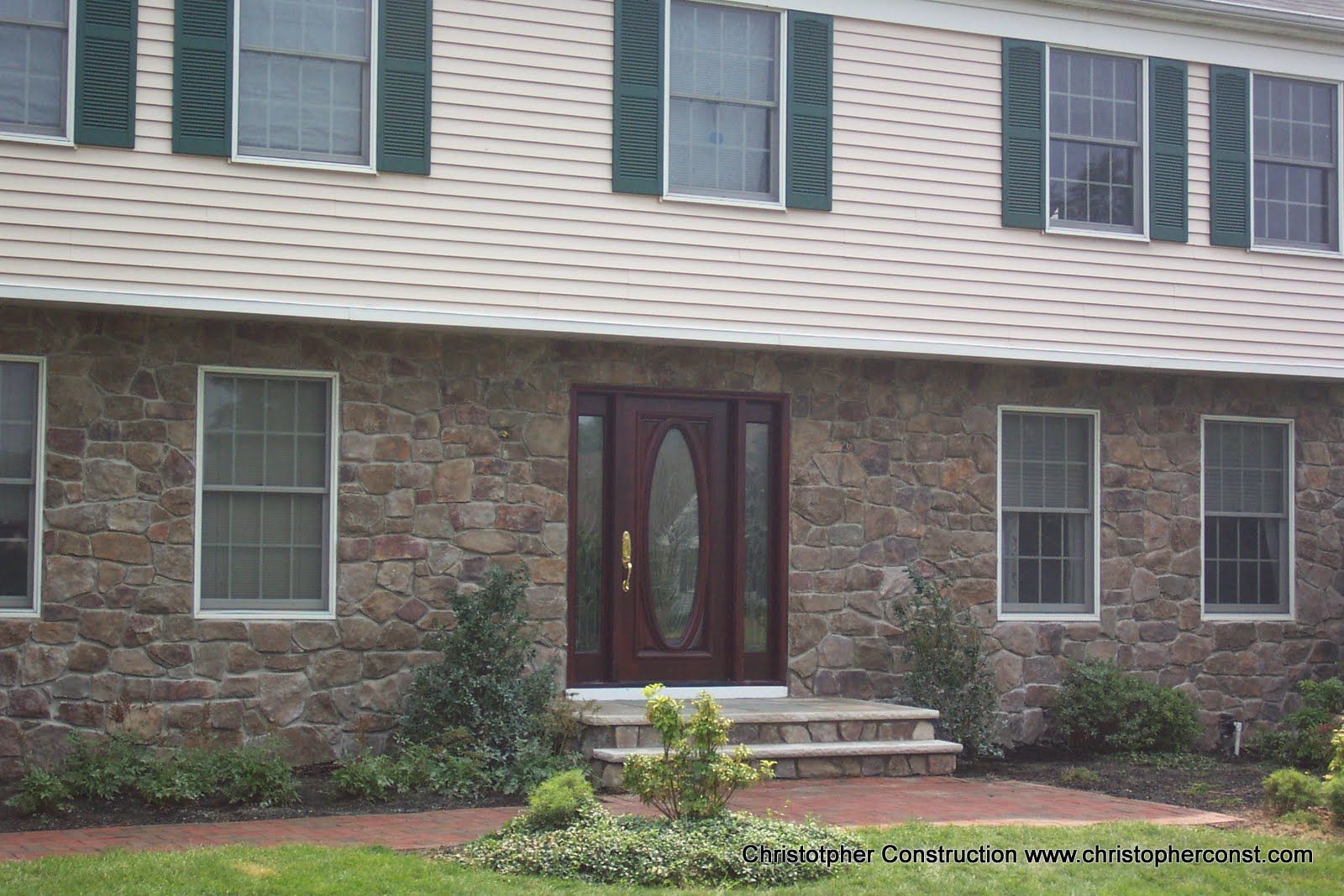Front entrance of a stone-and-siding house with a dark double door, steps, and small shrubs.