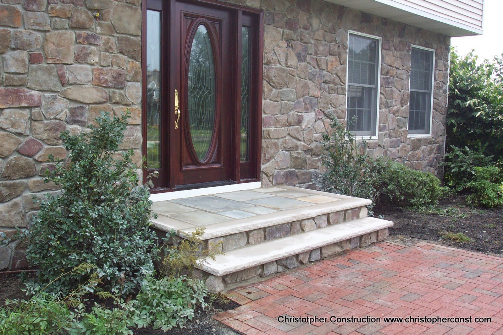 Front entrance of a stone house with steps, a red brick walkway, and shrubs on either side.