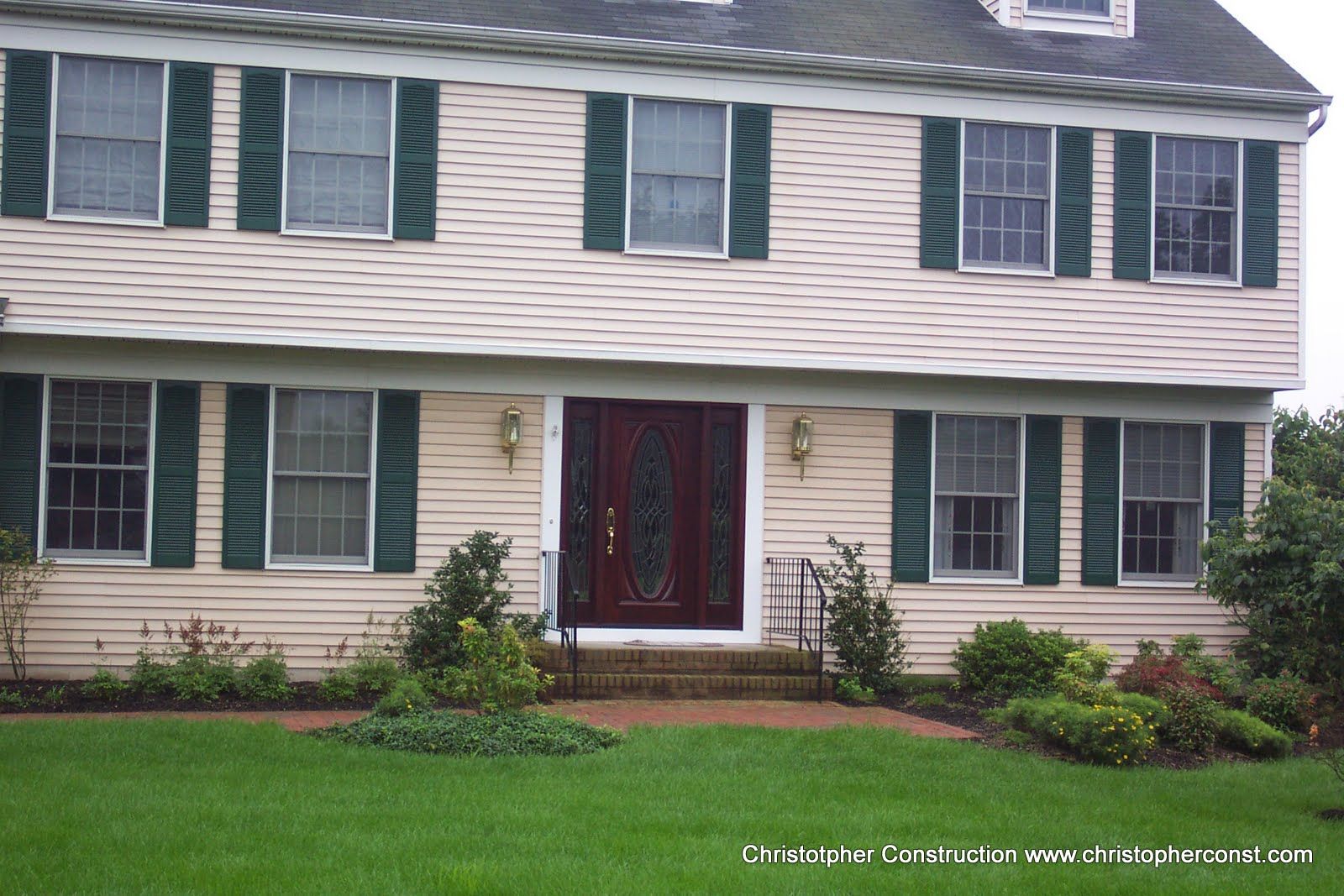 Two-story beige house with green shutters, red front door, and a small front garden