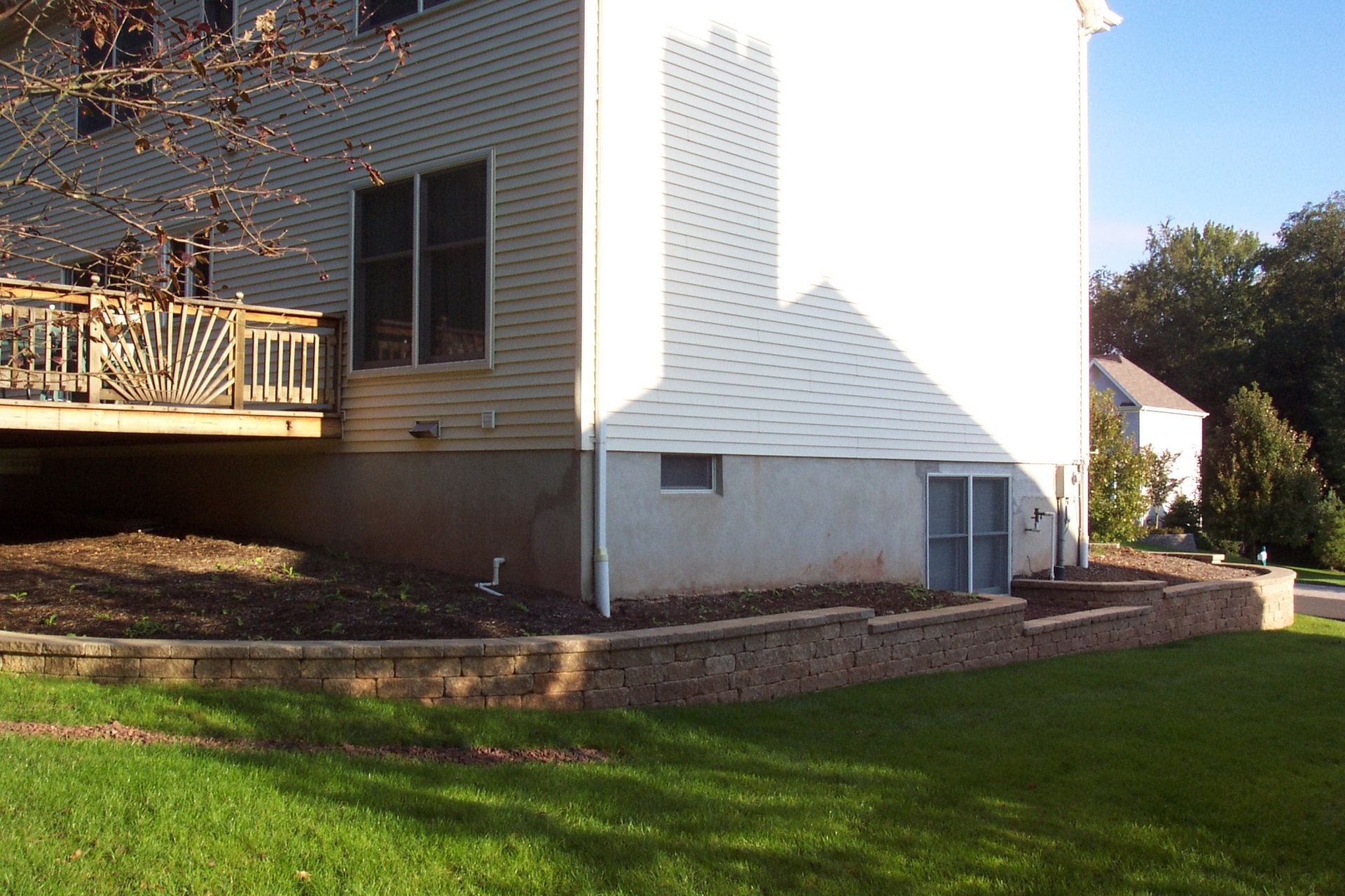 Suburban house with wooden deck, white siding, brick foundation, and green lawn in sunlight