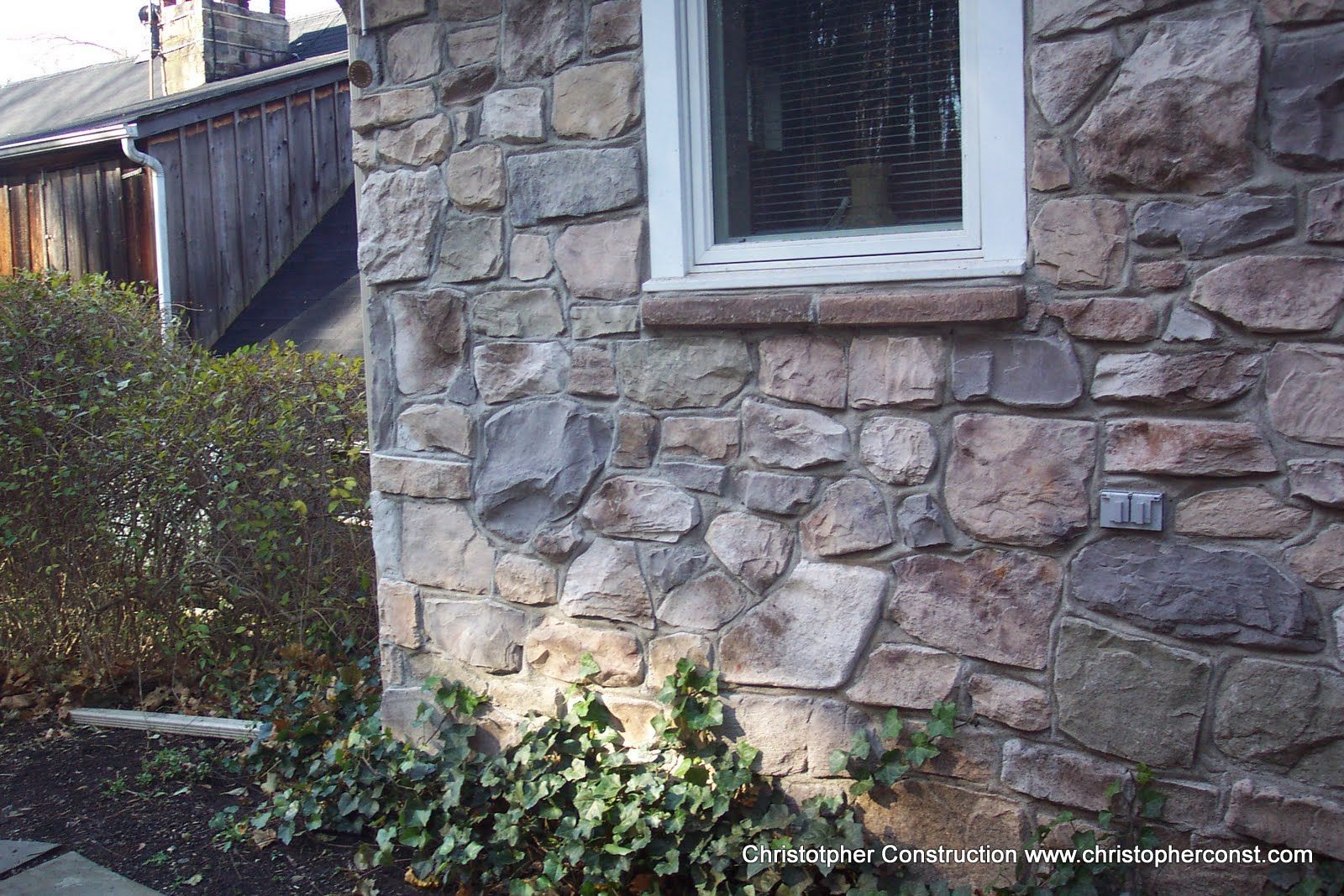 Stone house wall with a white-trimmed window beside shrubs and a wooden fence.