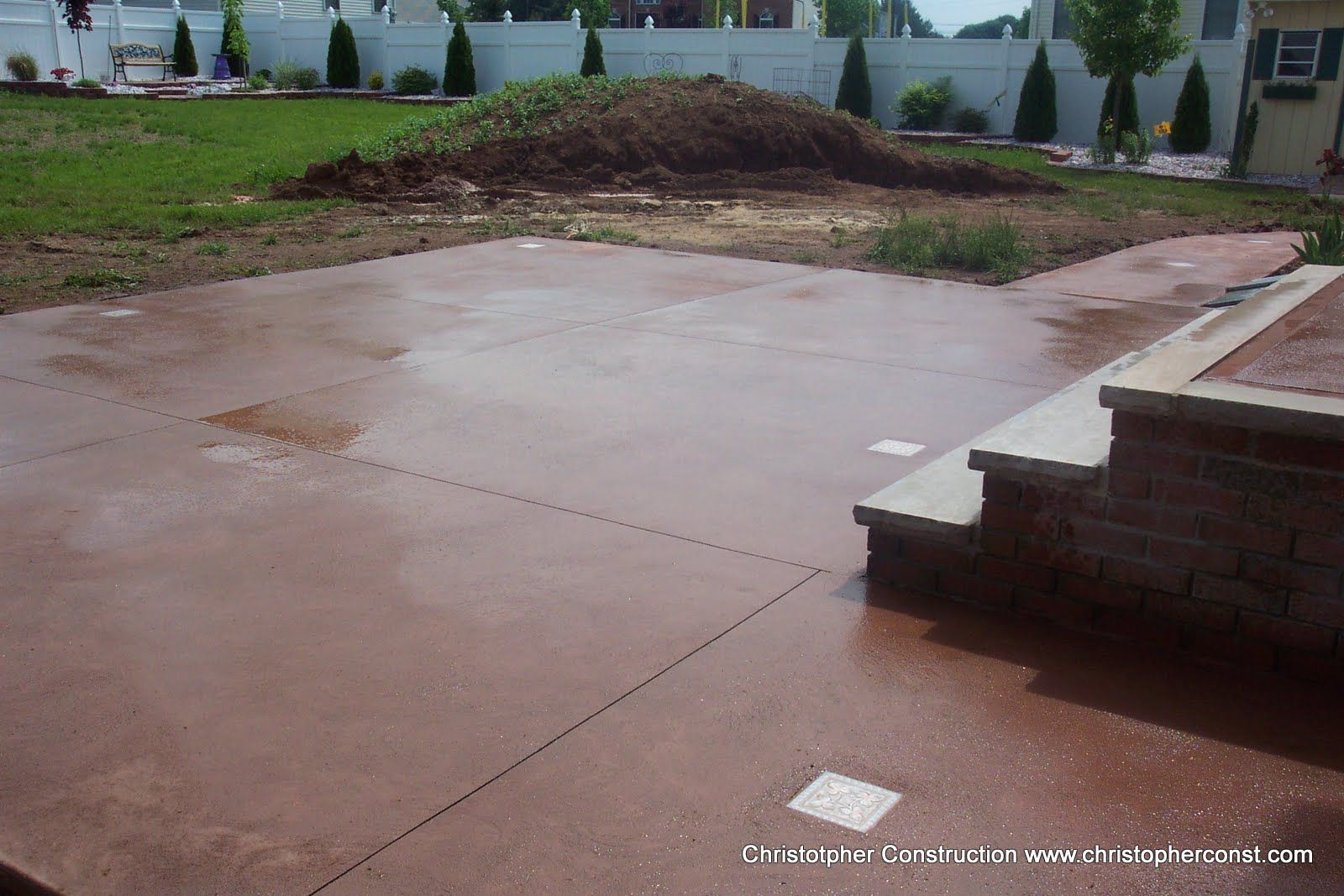 Freshly poured concrete patio in a backyard with a dirt pile and brick steps nearby