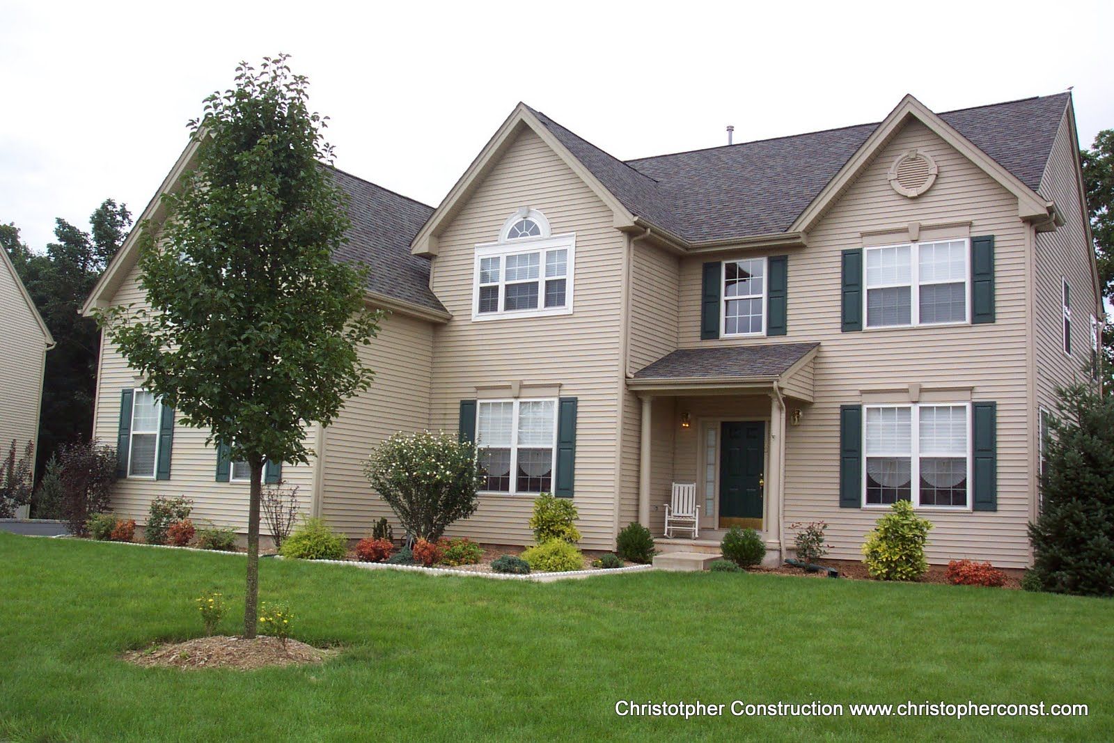 Large beige suburban house with stone accents, a front lawn, and a young tree in the yard
