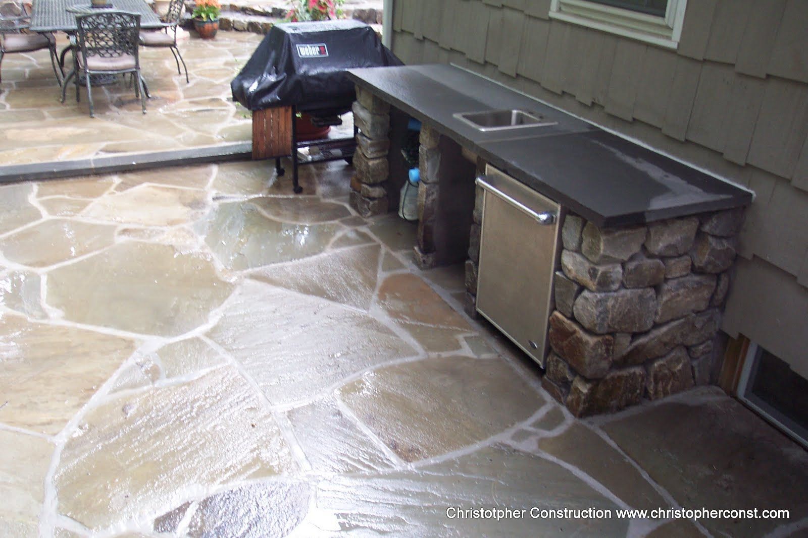 Outdoor stone patio with built-in grill and cabinets, wet from rain, beside a house