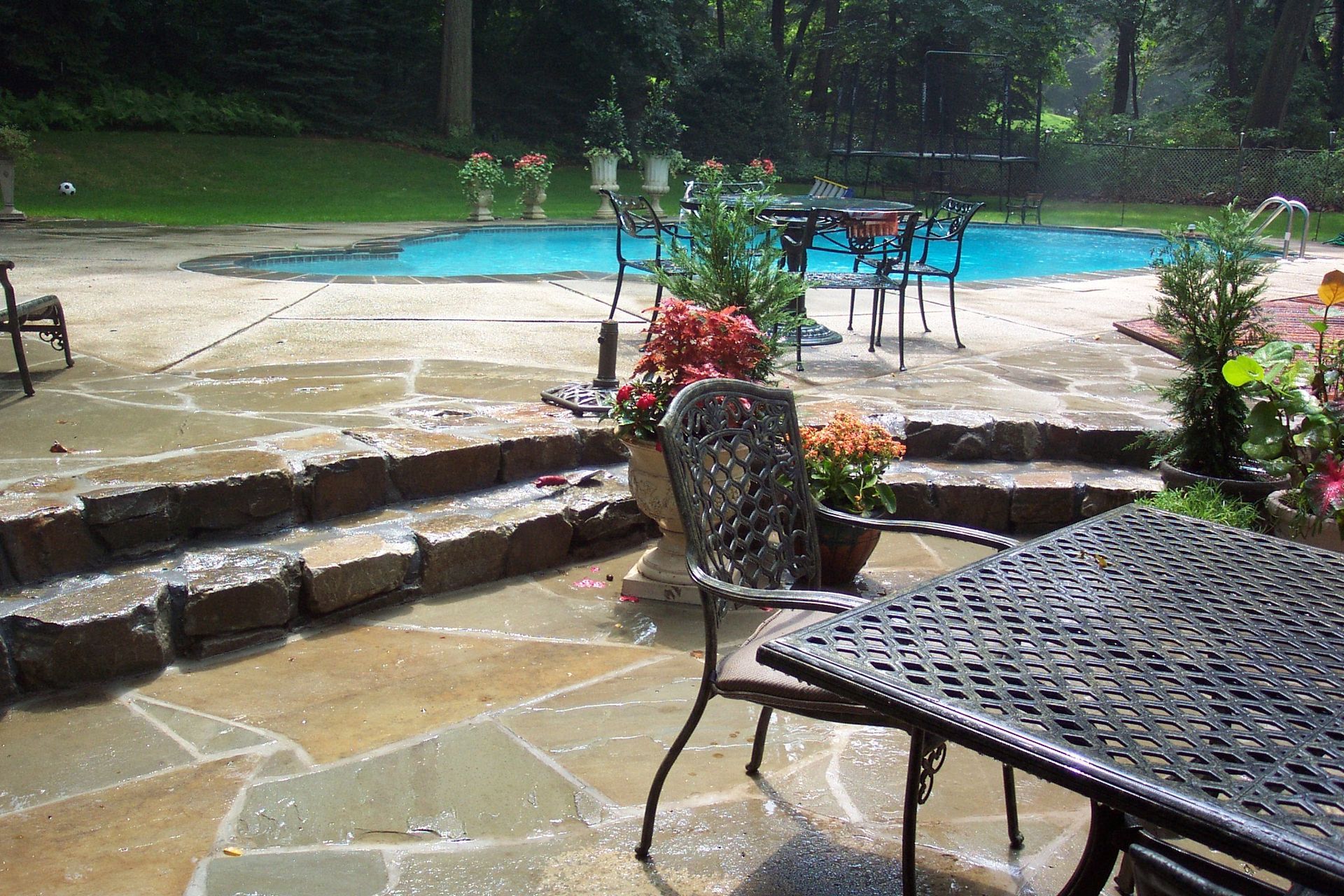 Stone patio with wet chairs and potted flowers beside a backyard swimming pool