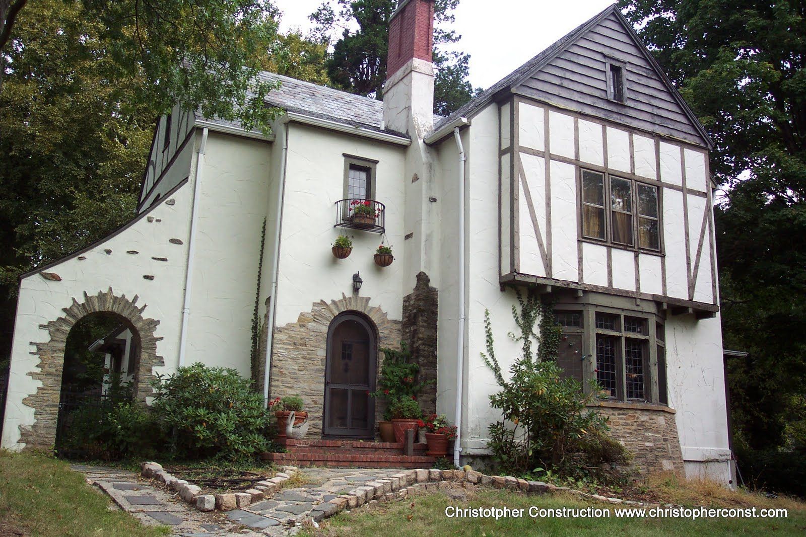 Small white cottage with dark trim, stone arch doorway, and ivy in a wooded setting