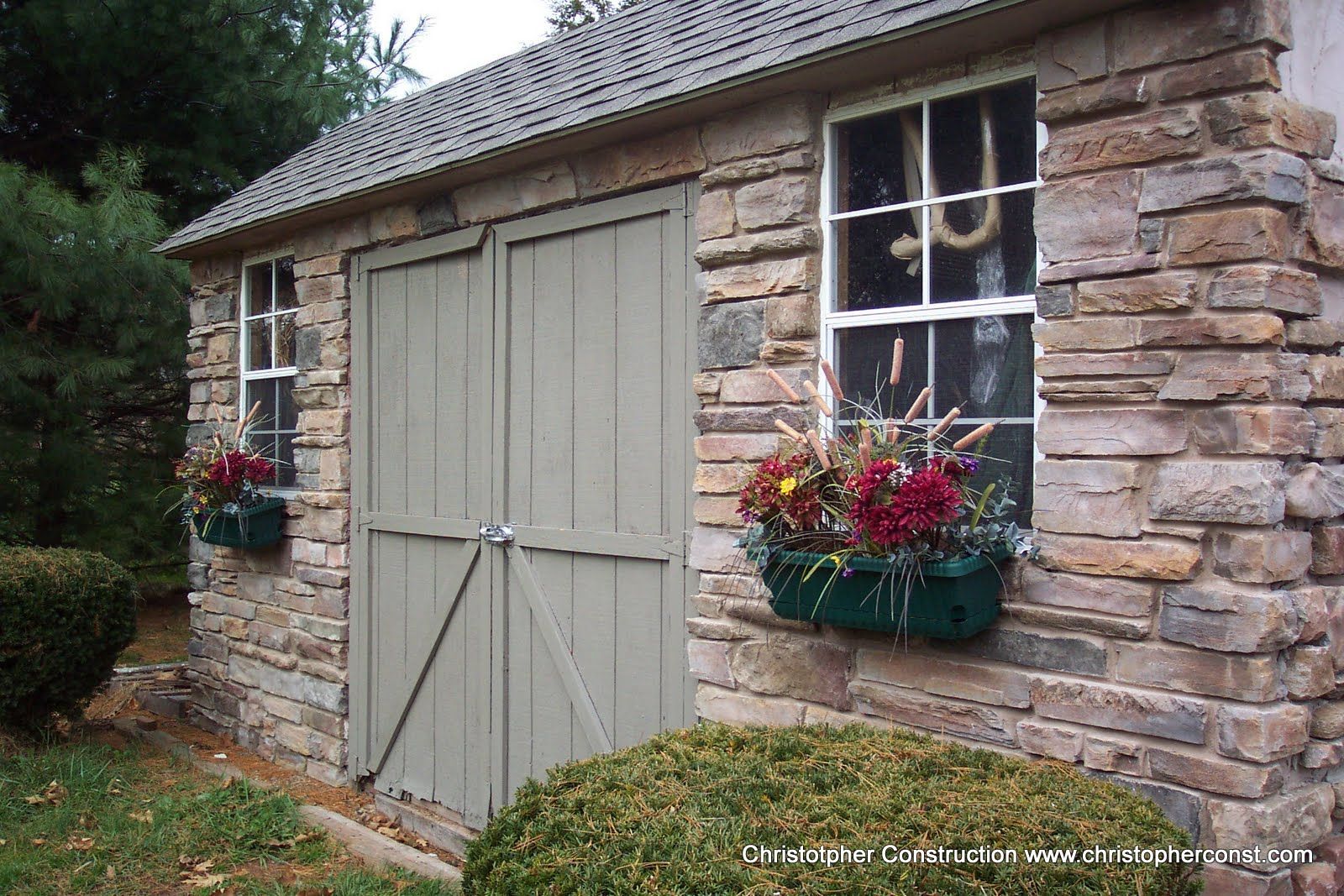 Stone garden shed with gray double doors, a window, and hanging flower baskets.
