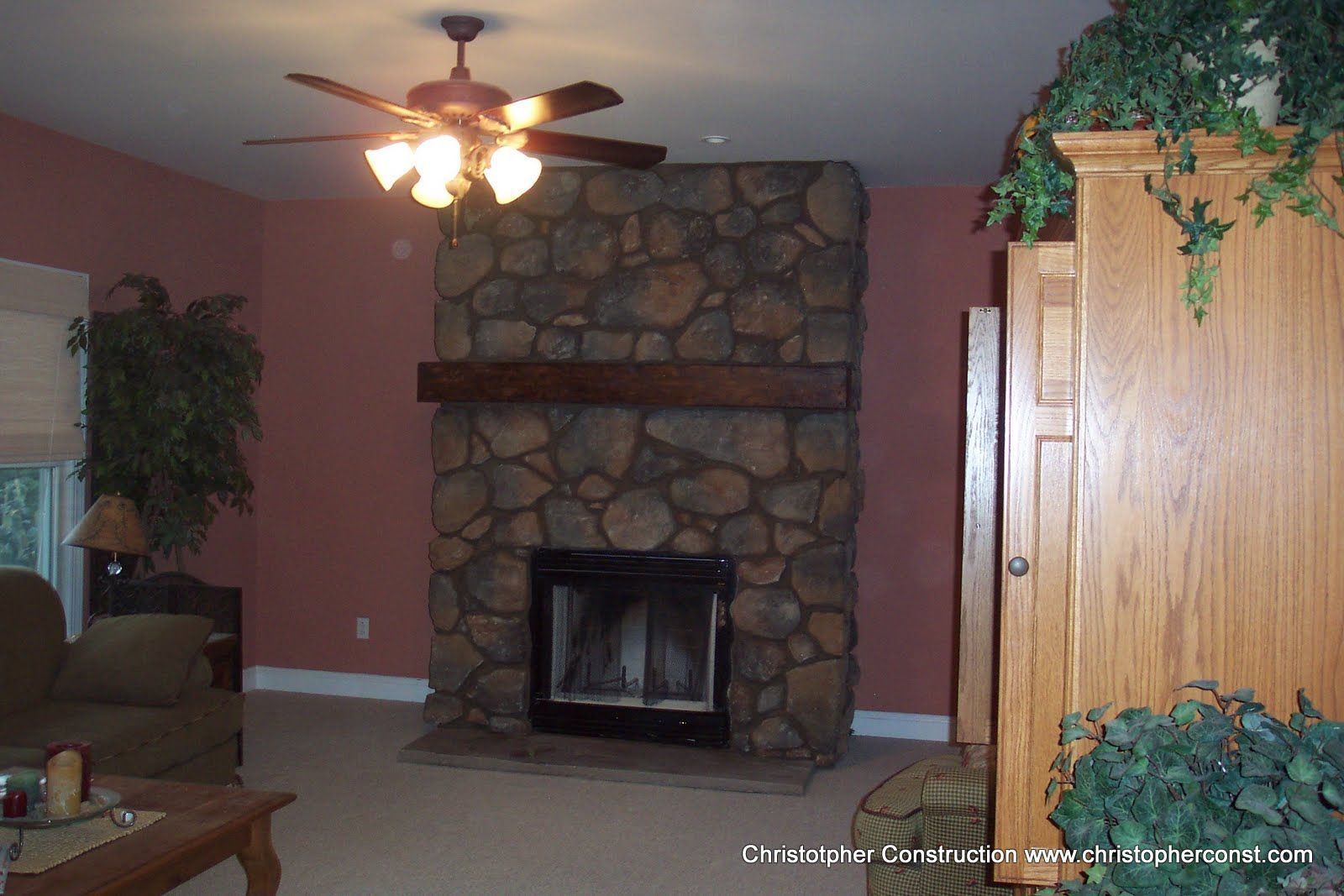 Living room with stone fireplace, ceiling fan, brown sofa, and wooden cabinet with hanging plants