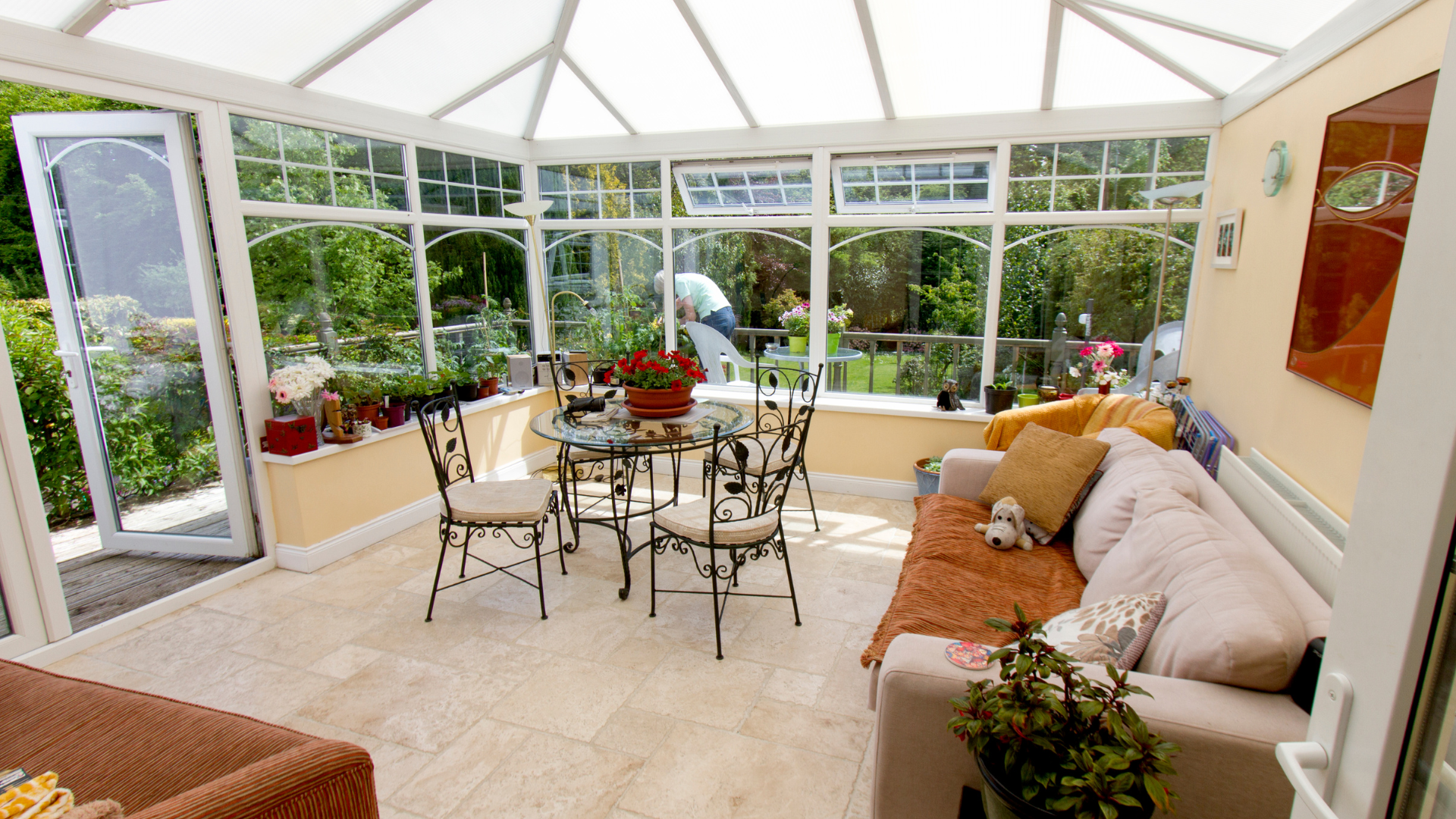 Bright sunroom with wicker seating, potted plants, and large windows overlooking a garden.