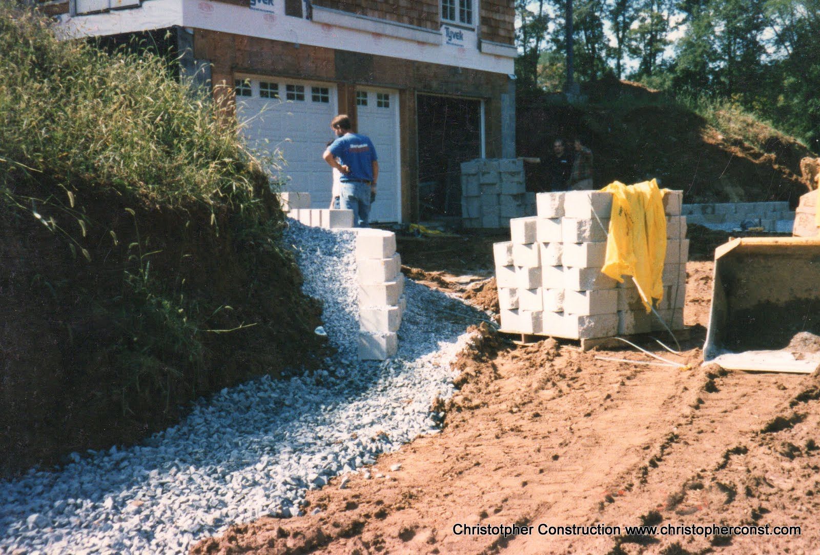 Worker laying gravel near a house foundation, with stacked blocks and a yellow tarp nearby