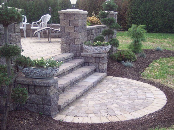 Backyard stone patio with steps, curved paver walkway, and raised garden planters