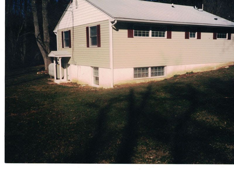 Small beige two-story house with white trim on a grassy lot, viewed from the side in sunlight.