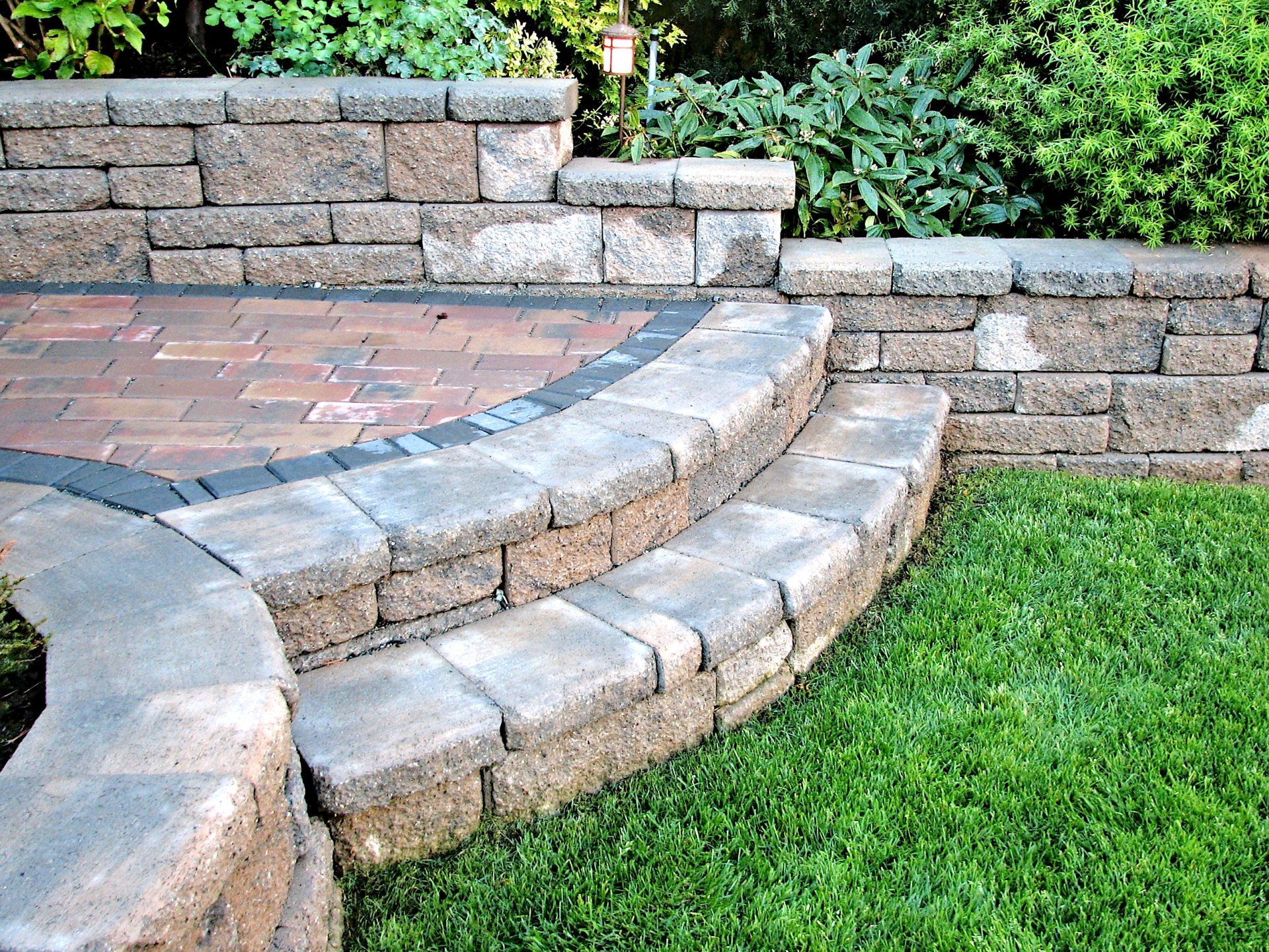 Curved stone steps beside a brick patio and green lawn, with a low retaining wall and shrubs.