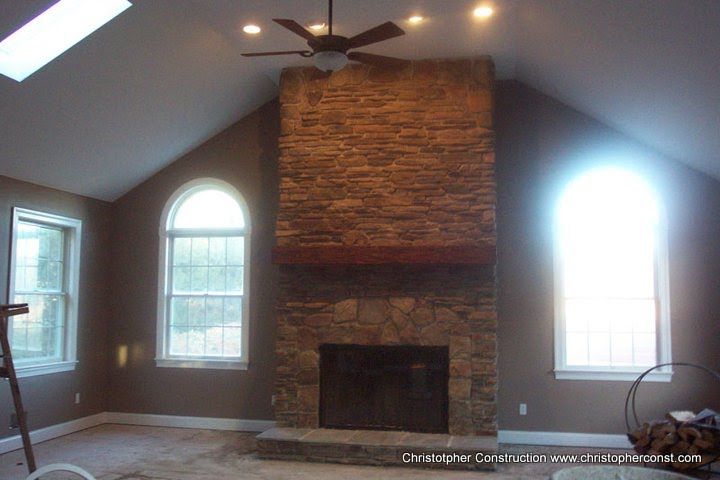 Living room with vaulted ceiling, stone fireplace, and arched windows, lit by a ceiling fan and natural light