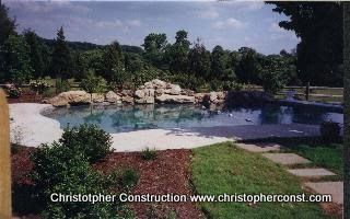 Backyard pool with rock waterfall, trees, and a stone patio under a cloudy sky