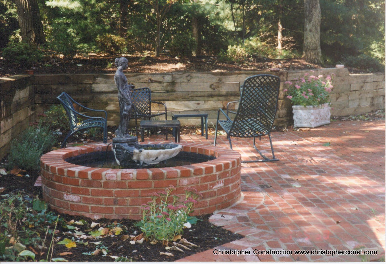 Brick fountain in a brick patio garden with two metal chairs and potted flowers
