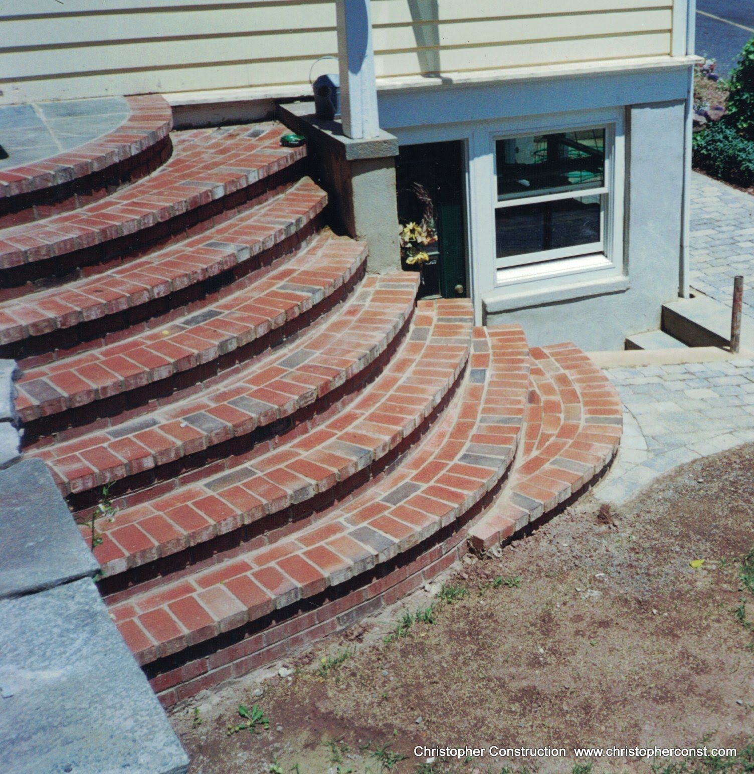 Curved red brick steps leading to a house entrance beside a basement window and beige siding