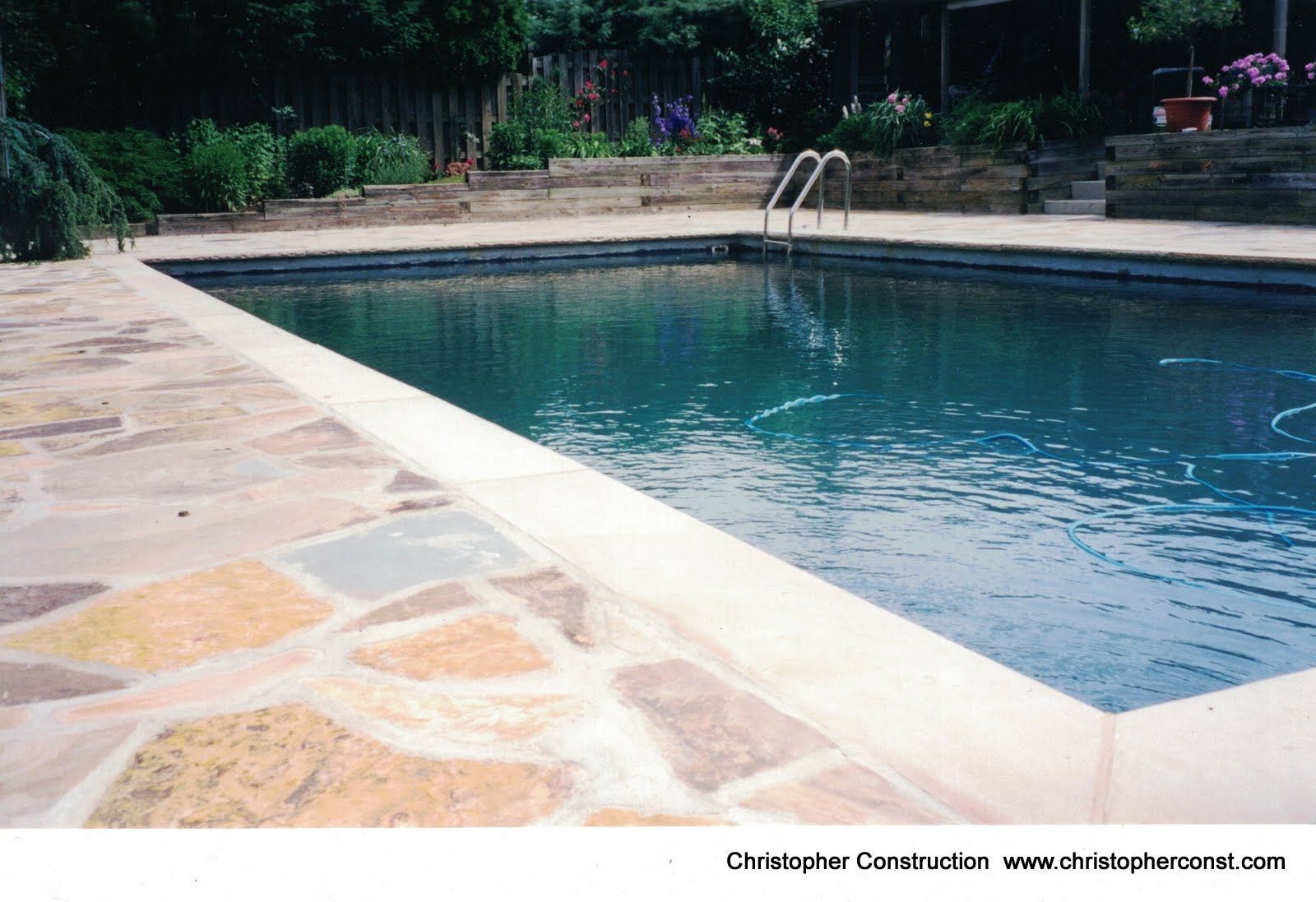 Swimming pool with clear blue water beside a stone deck and ladder, surrounded by greenery.