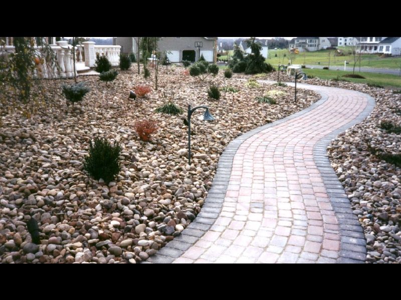Curving brick garden path through a rocky, mulched landscape with small shrubs and homes in the background