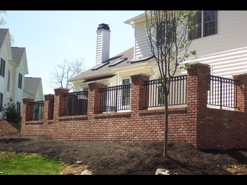 Brick house with a fenced front yard and a young tree in a landscaped suburban neighborhood.