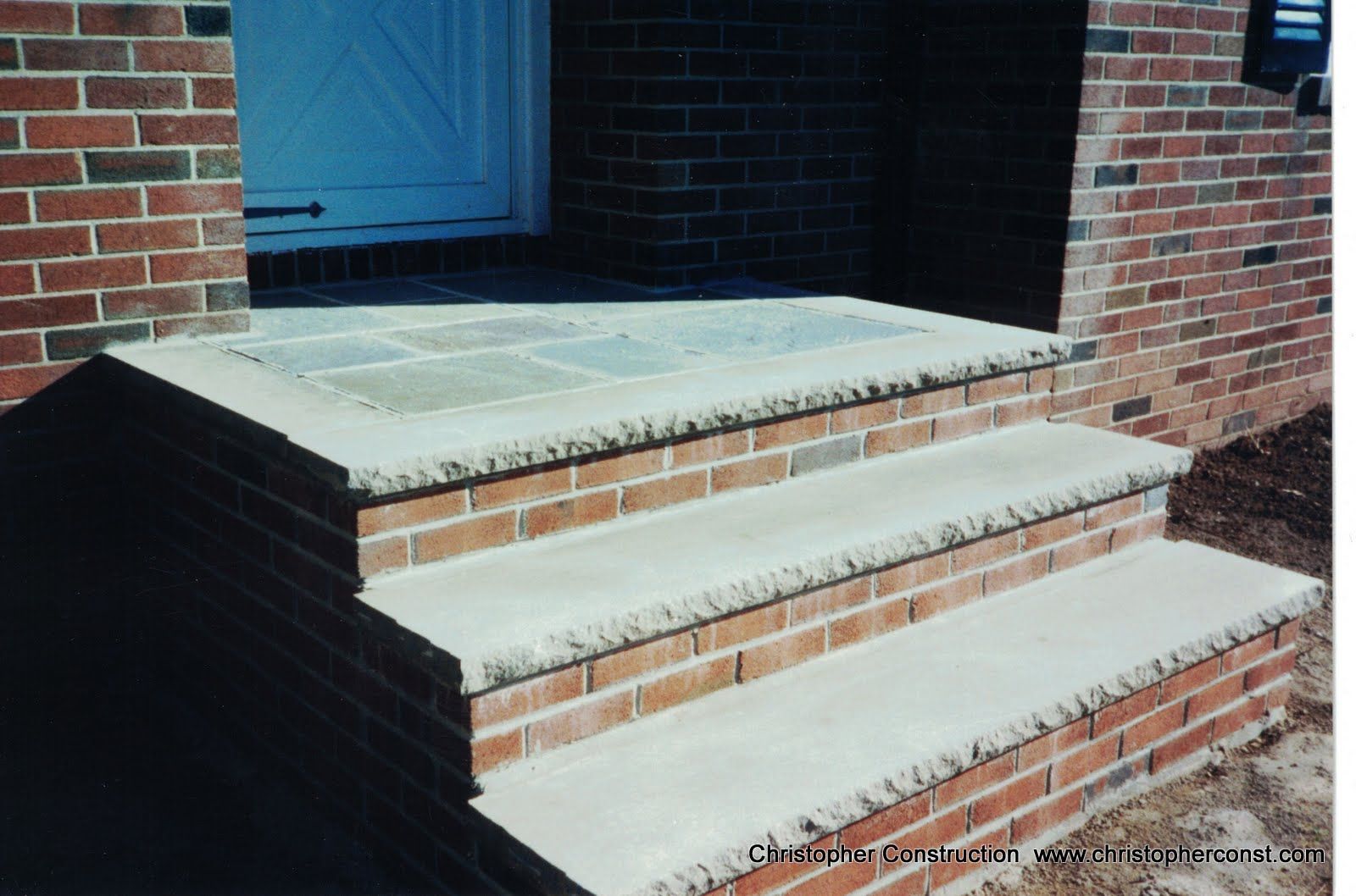 Brick front steps leading to a porch with a blue door and railing handle