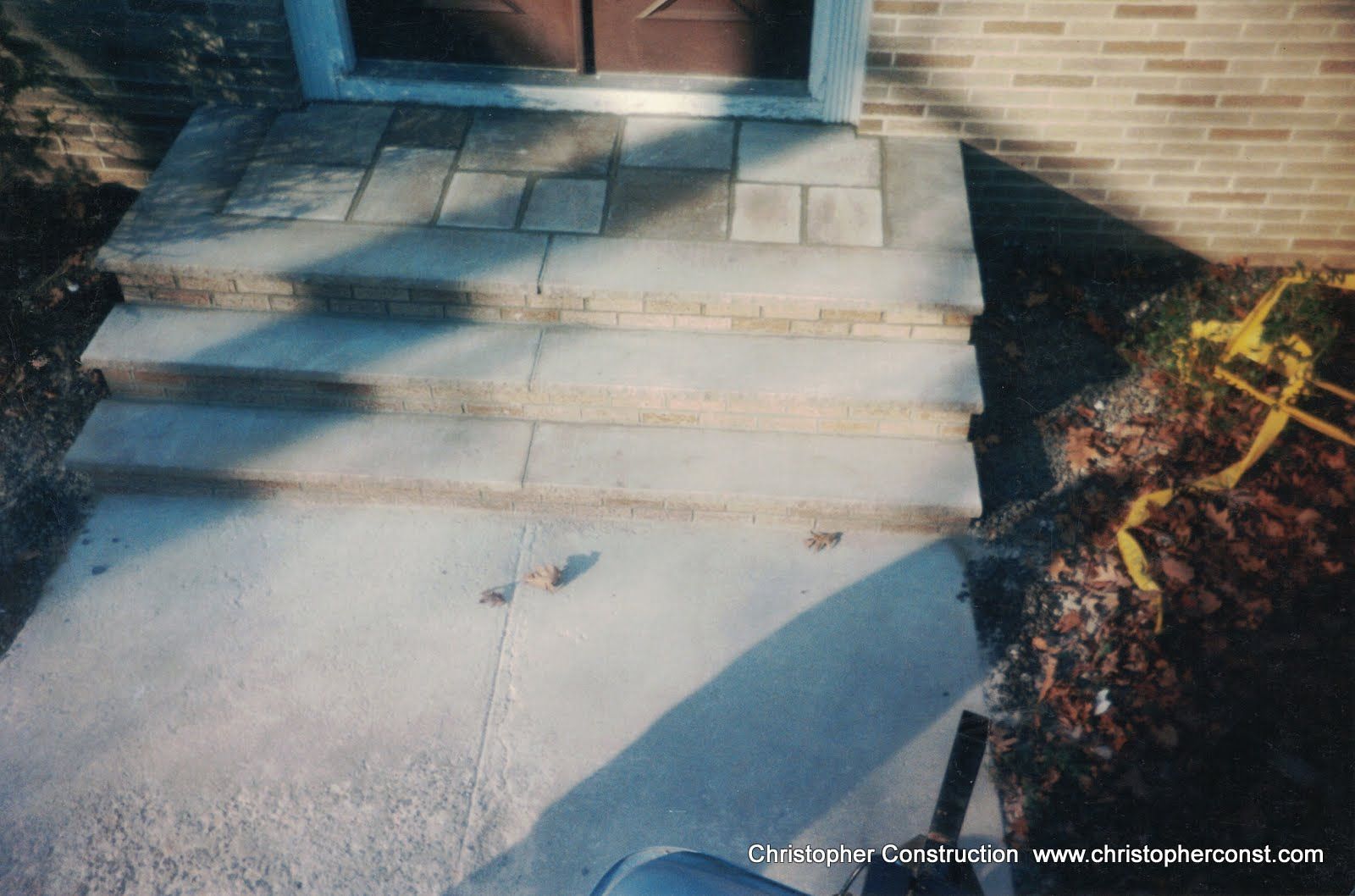Front steps and walkway leading to a brick house entrance, with a concrete path and flower bed.