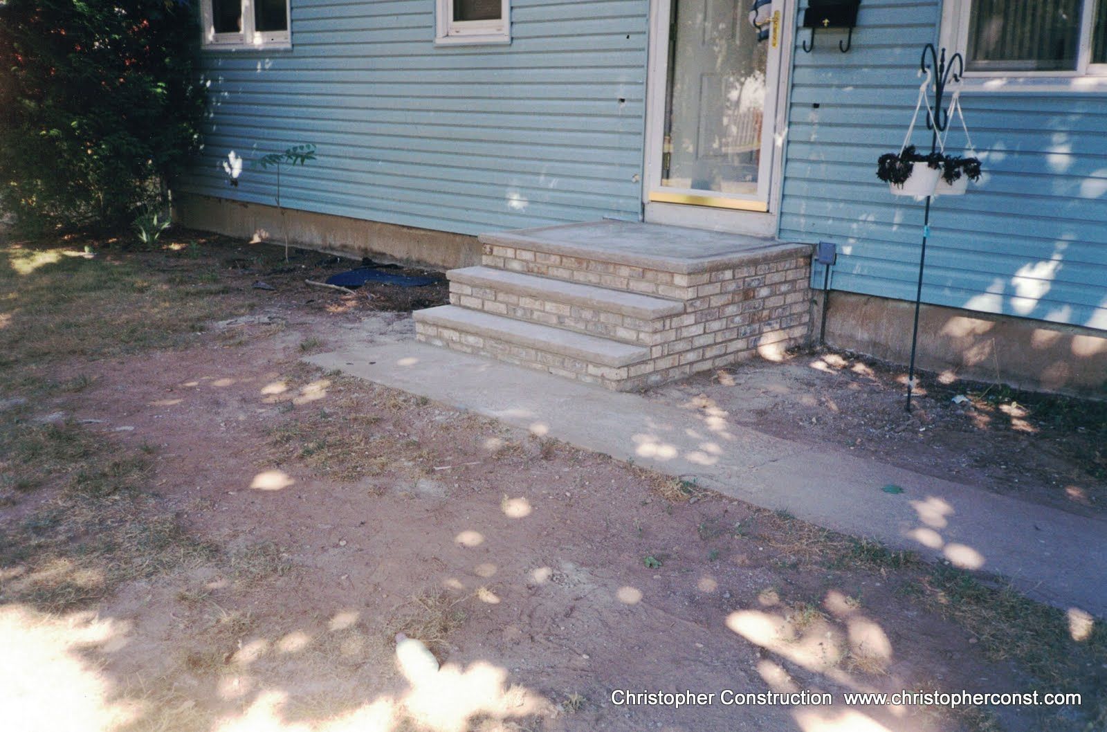 Blue house exterior with concrete steps, small porch, and shaded dirt yard