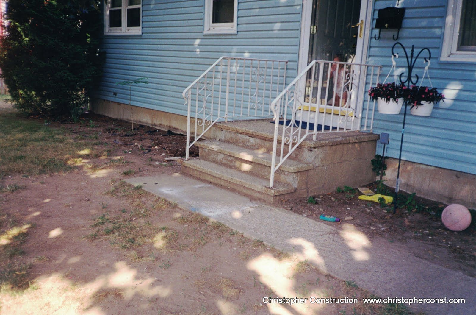 Front steps of a blue house with white railing, concrete walkway, hanging flowers, and a pink ball in the yard
