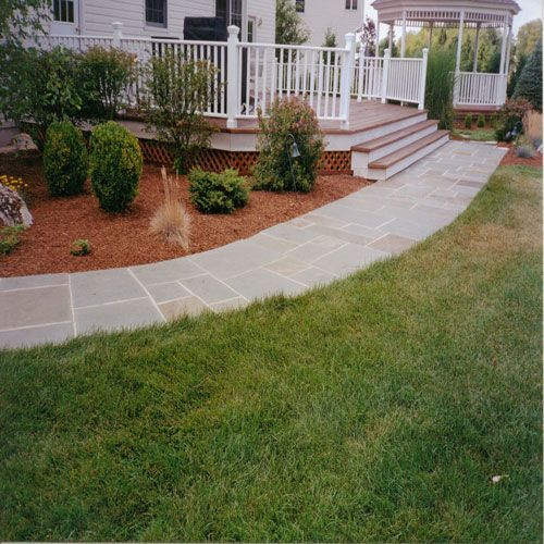 Curved stone walkway beside a raised deck with white railing and shrubs, bordered by green lawn.