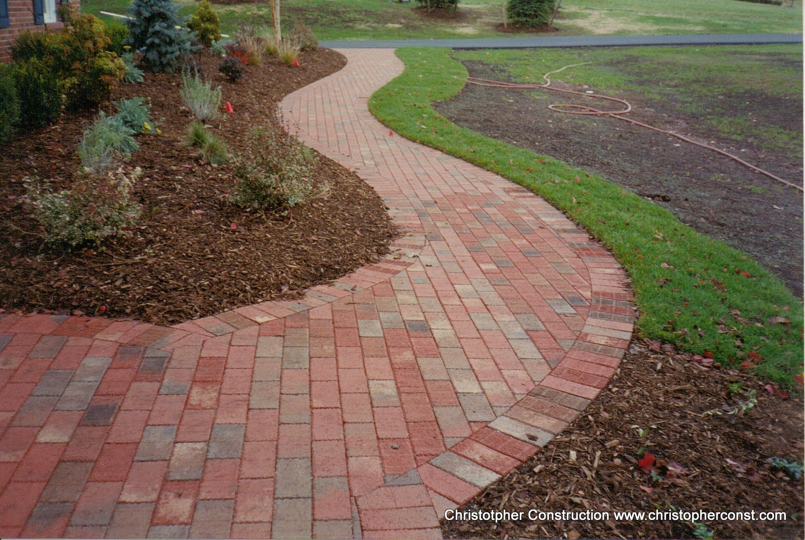Curving brick garden path bordered by mulch, grass, and shrubs in a landscaped yard
