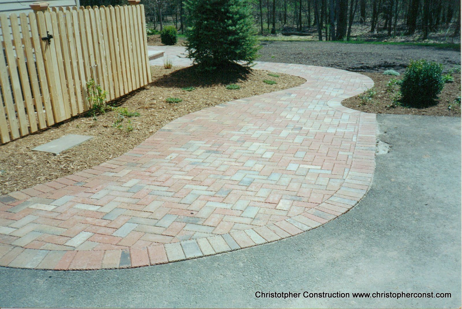 Curved brick garden walkway beside a wooden fence and shrubs in a landscaped yard