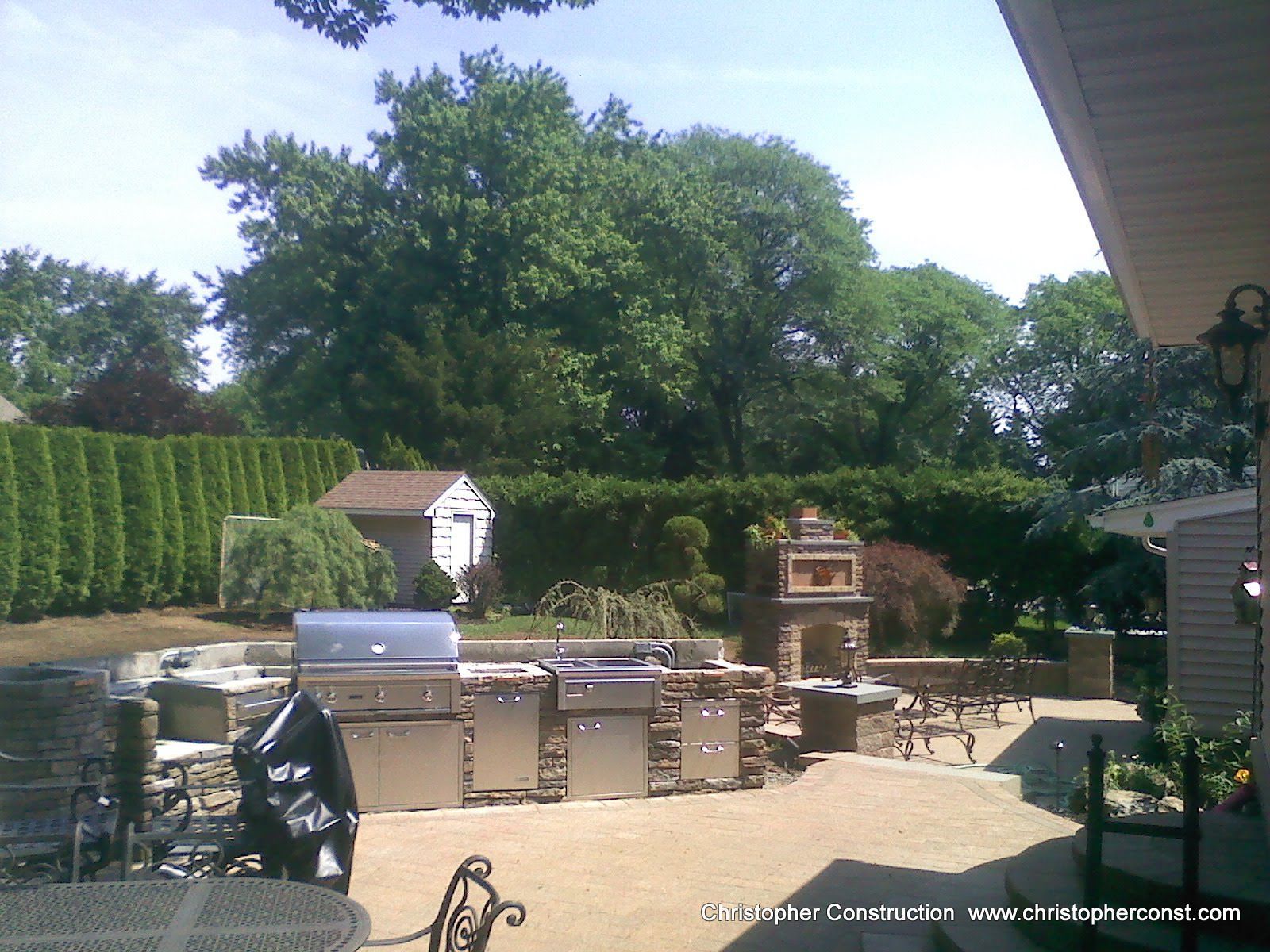 Backyard patio with outdoor grill, metal counters, and a brick oven under trees and a sunny sky