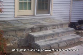 Stone front steps and landing leading to a house door with gray siding and a window