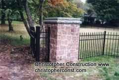 Stone gate pillar beside black metal fence in a grassy yard with trees.
