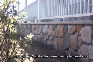 Stone retaining wall beneath a light gray fence beside a landscaped slope