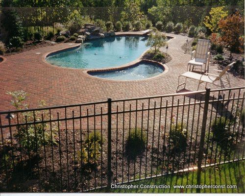 Backyard pool with brick patio, black metal fence, and lounge chair in a landscaped yard