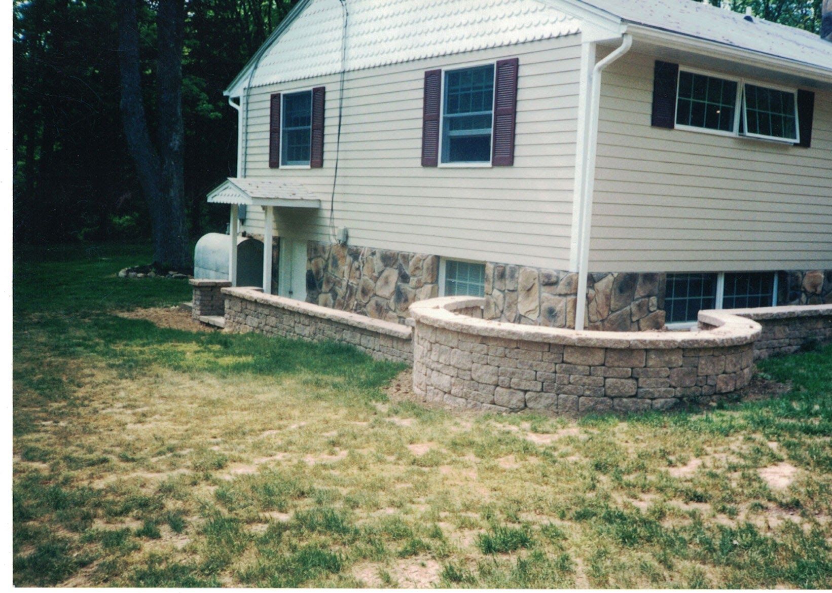 Two-story beige house with stone foundation and curved retaining wall on a grassy yard