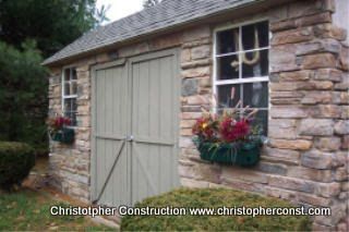 Stone garden shed with double wooden doors, window boxes of red flowers, and a pitched roof