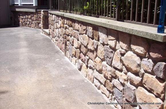 Stone retaining wall along a curved concrete walkway beside a metal fence in bright sunlight
