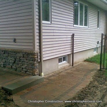 Side view of a house with vinyl siding, stone foundation, and a wet brick walkway with metal handrails