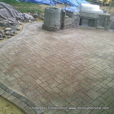 Brick-paved patio with curved edge beside stone outdoor kitchen and rocky hillside.
